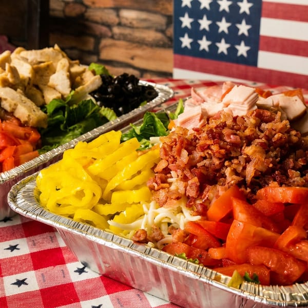 Two catering trays of salads with various toppings on a red and white checkered tablecloth.