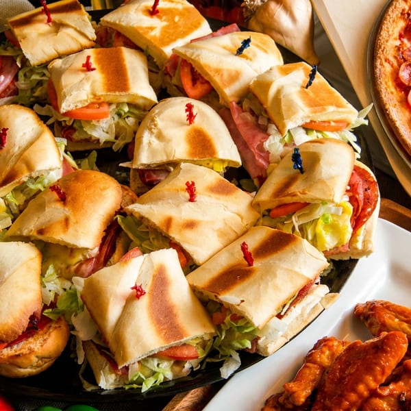 Platter of small subs, a pepperoni pizza, and buffalo chicken wings on a table.