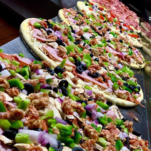 Rows of uncooked pizzas with various toppings like ham, sausage, peppers, onions, and olives.